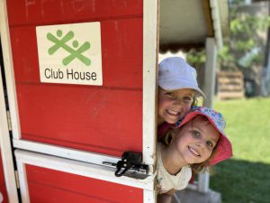 Two children peeking around the corner of a red door with the BGC Okanagan logo on it. BGC Okanagan will continue Child Care Referral and Resource services in the North Okanagan.